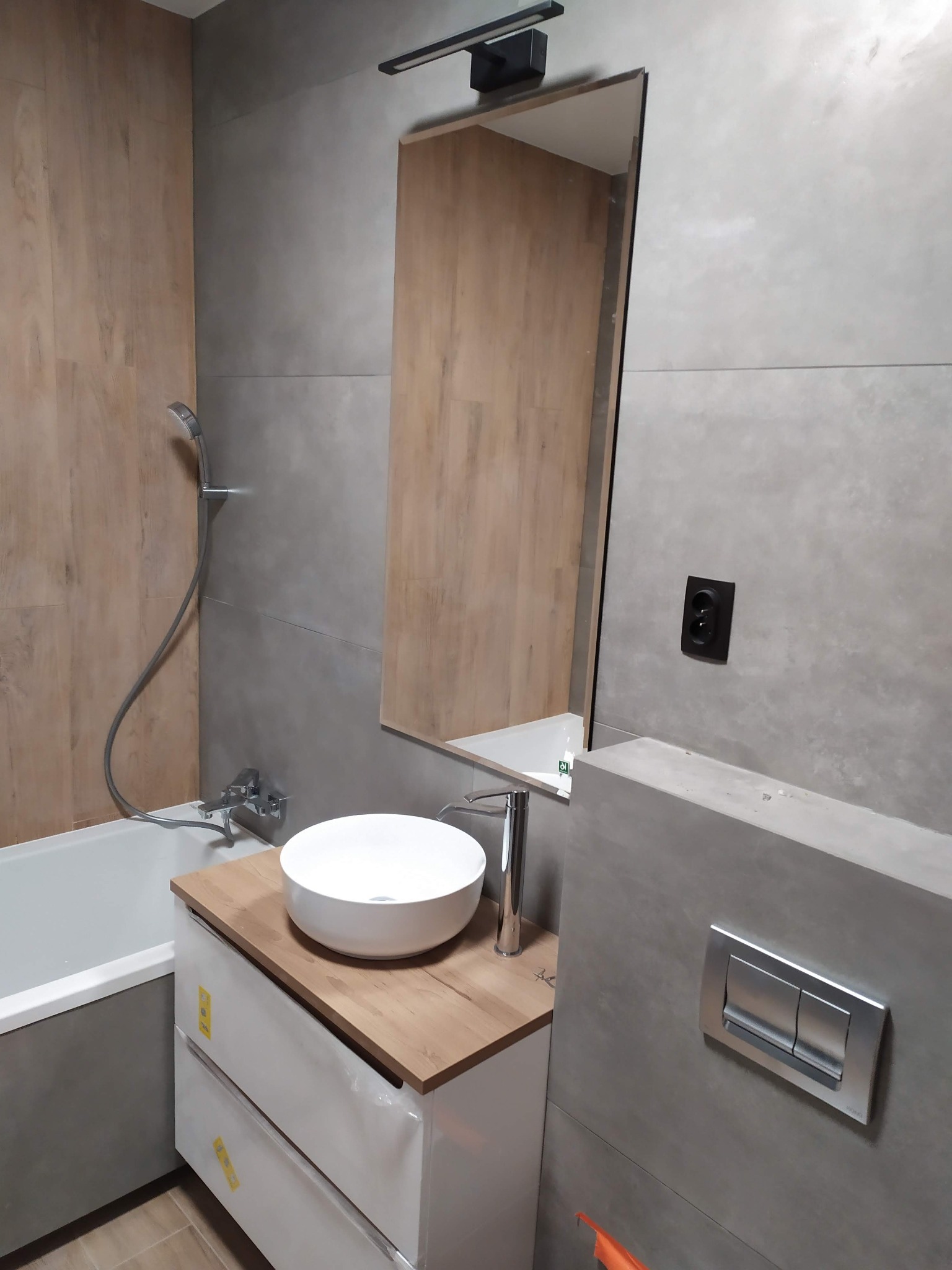 Nowoczesna łazienka z okrągłą umywalką nablatową, drewnianym blatem i szarą armaturą, widoczny fragment wanny i lustro z oświetleniem.