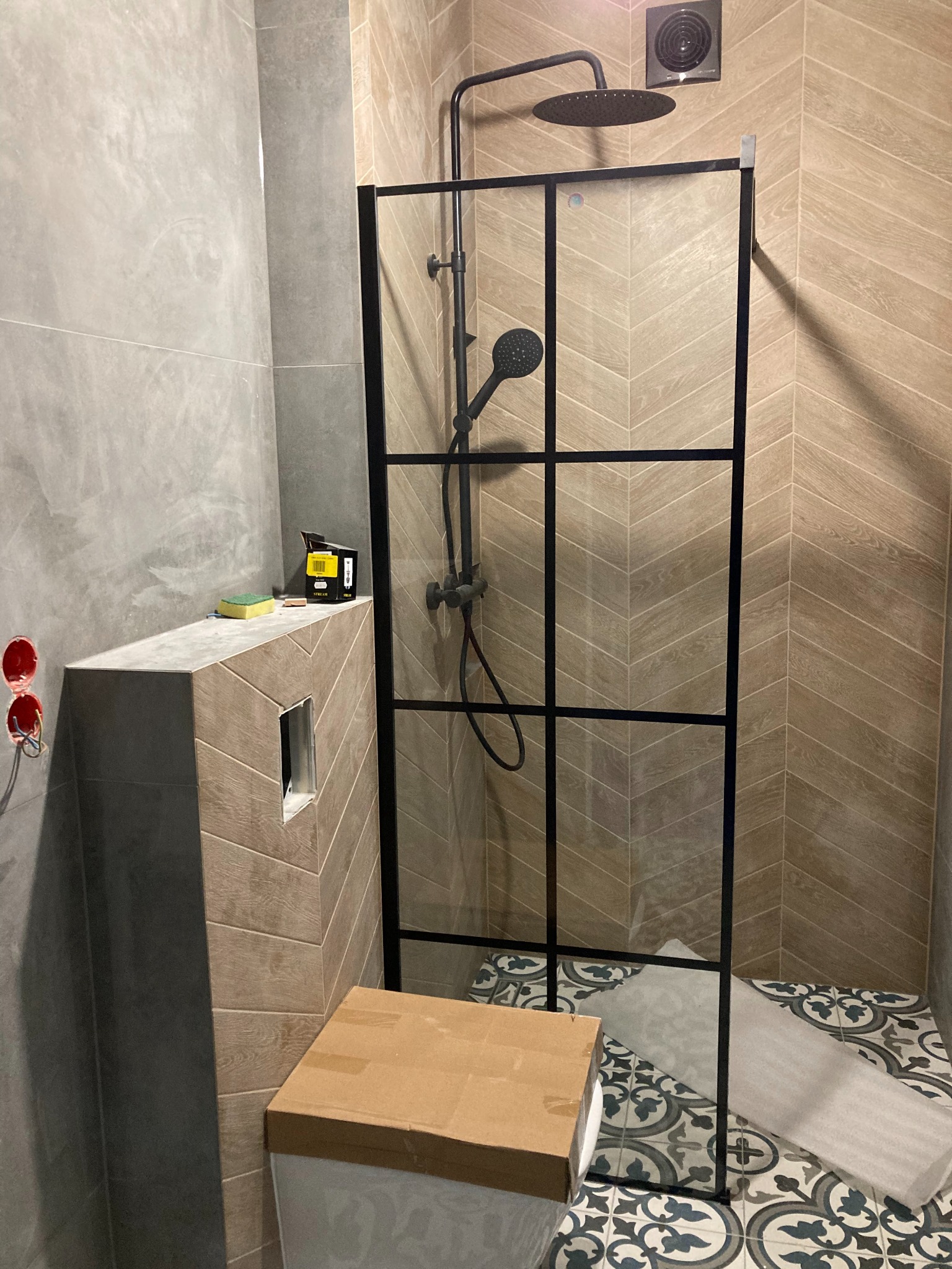 Montaż kabiny prysznicowej typu loft z czarnymi profilami, widoczne płytki imitujące drewno ułożone w jodełkę na ścianie oraz ozdobne płytki podłogowe, częściowo zabezpieczone folią ochronną.