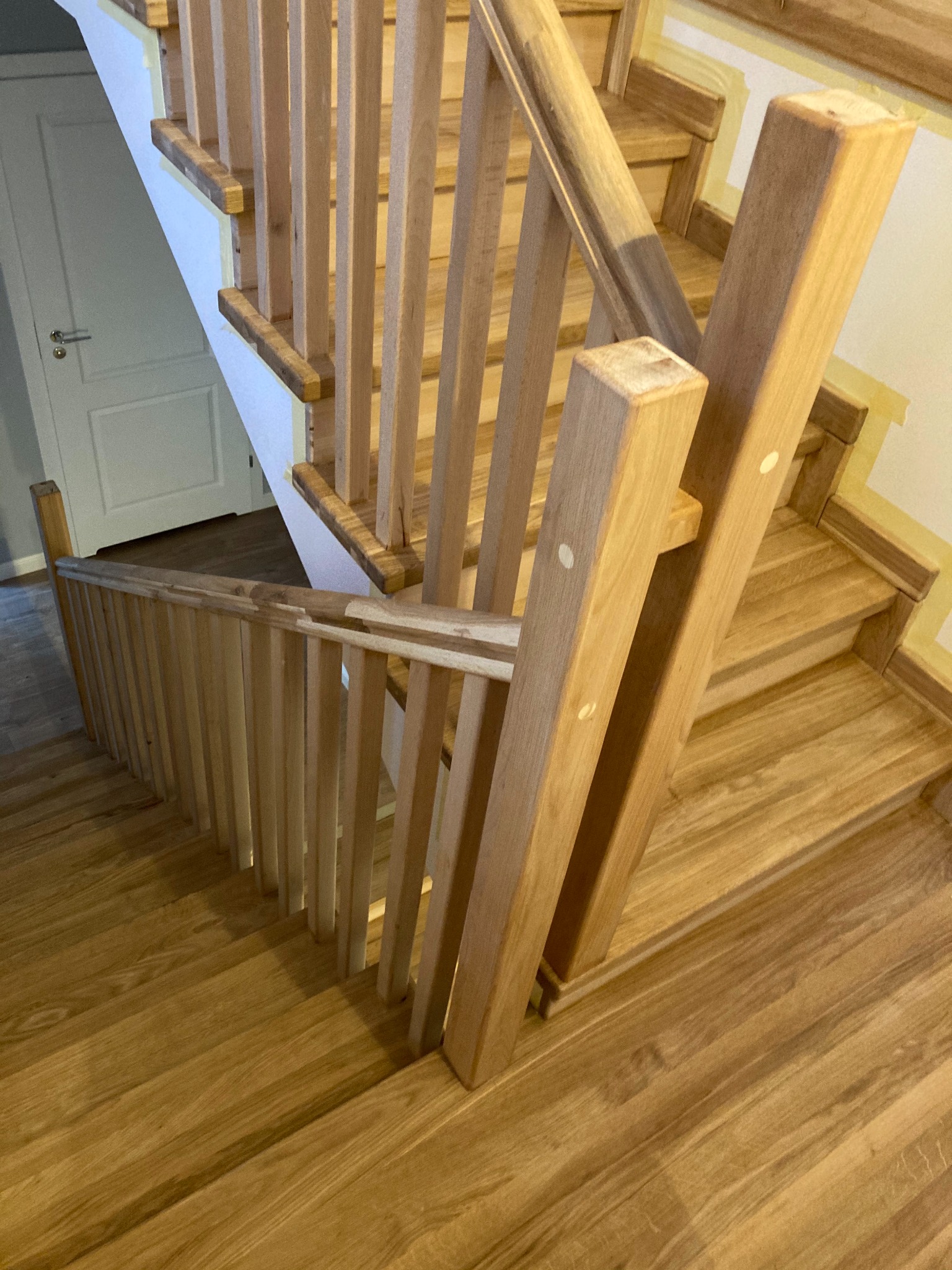 Drewniane schody z balustradą w trakcie montażu, zabezpieczone taśmą malarską przy ścianie, widok z góry na stopnie i poręcz.