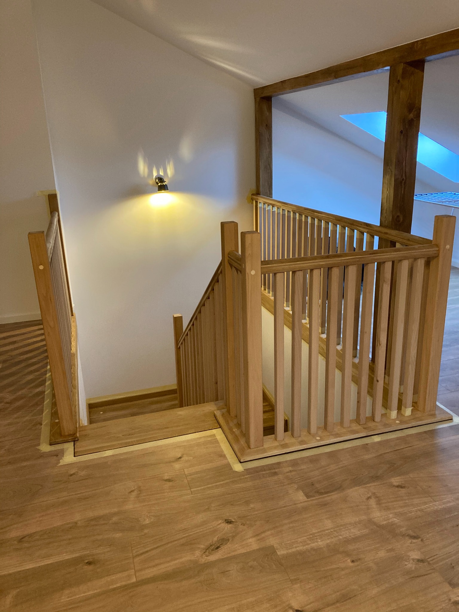 Drewniane schody z balustradą w jasnym odcieniu drewna, widoczne taśmy malarskie zabezpieczające podłogę, oświetlenie punktowe na ścianie, fragment konstrukcji drewnianej poddasza.