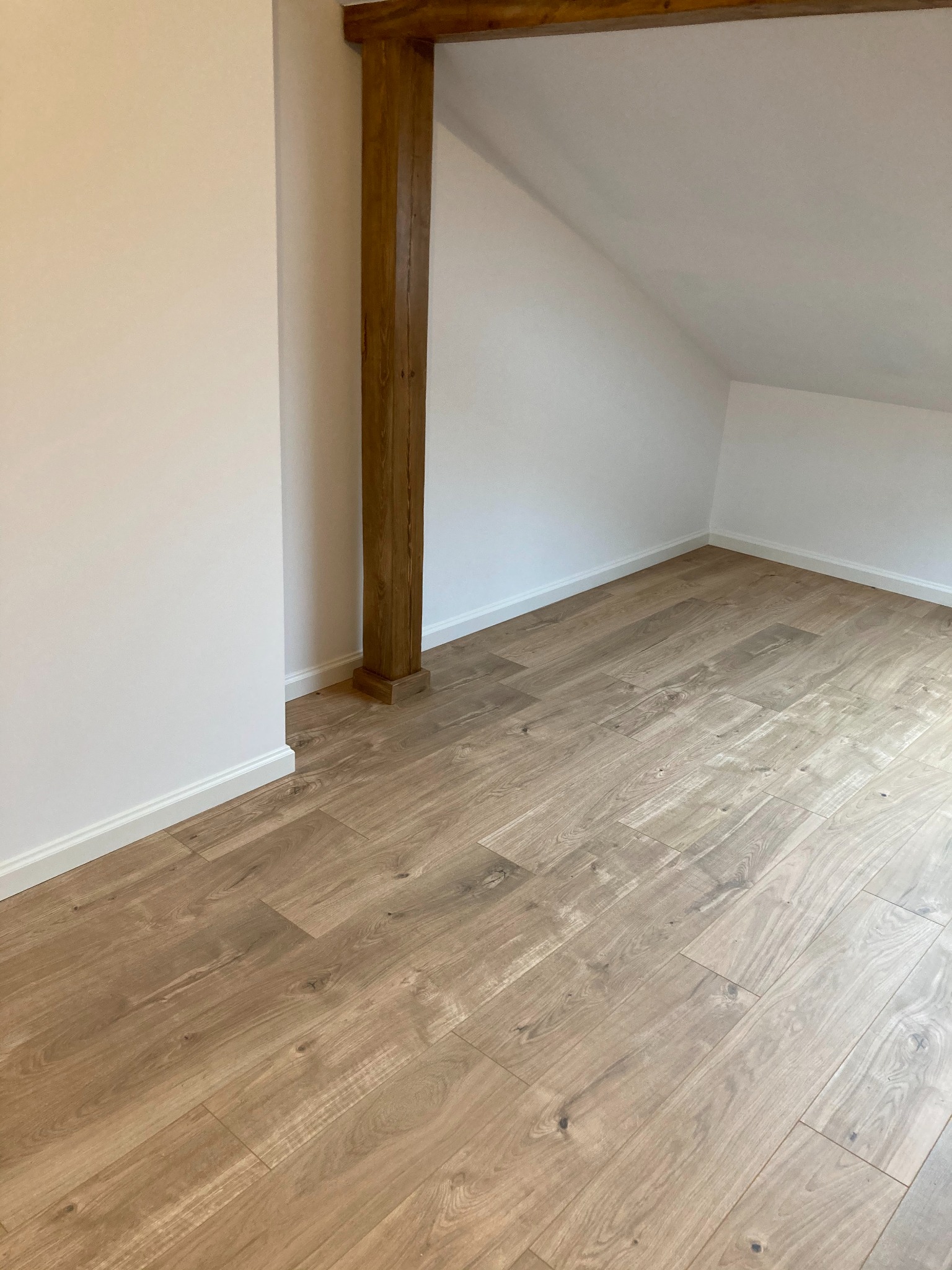 Poddasze po remoncie: świeżo położona podłoga z paneli imitujących jasne drewno, białe ściany i listwy przypodłogowe, widoczna drewniana belka konstrukcyjna.