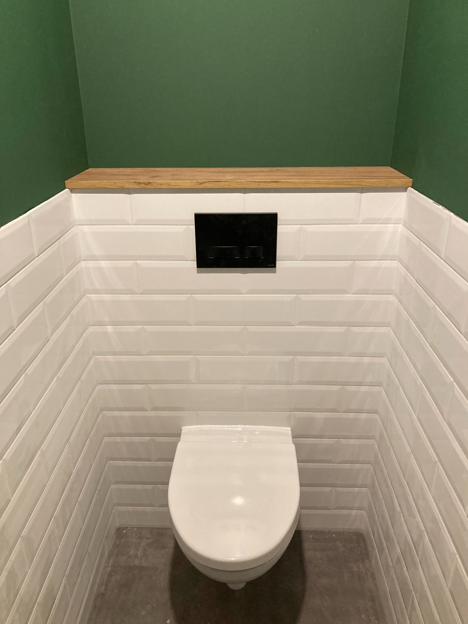 Nowoczesna toaleta wnękowa z białymi, prostokątnymi płytkami ułożonymi w cegiełkę, drewnianą półką nad spłuczką i zieloną ścianą w tle.