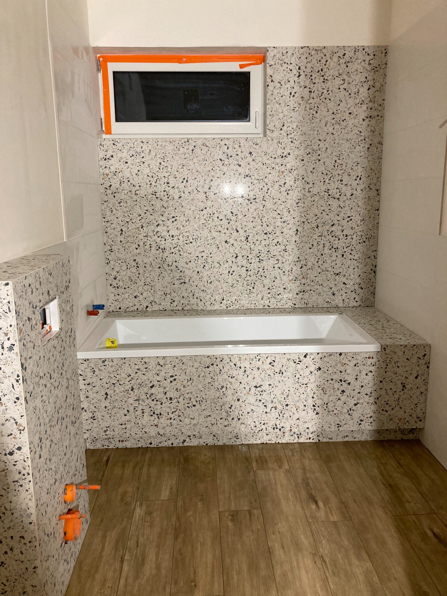 Nowoczesna łazienka z wanną obudowaną płytkami lastryko, drewnopodobna podłoga i okno z pomarańczowym zabezpieczeniem.