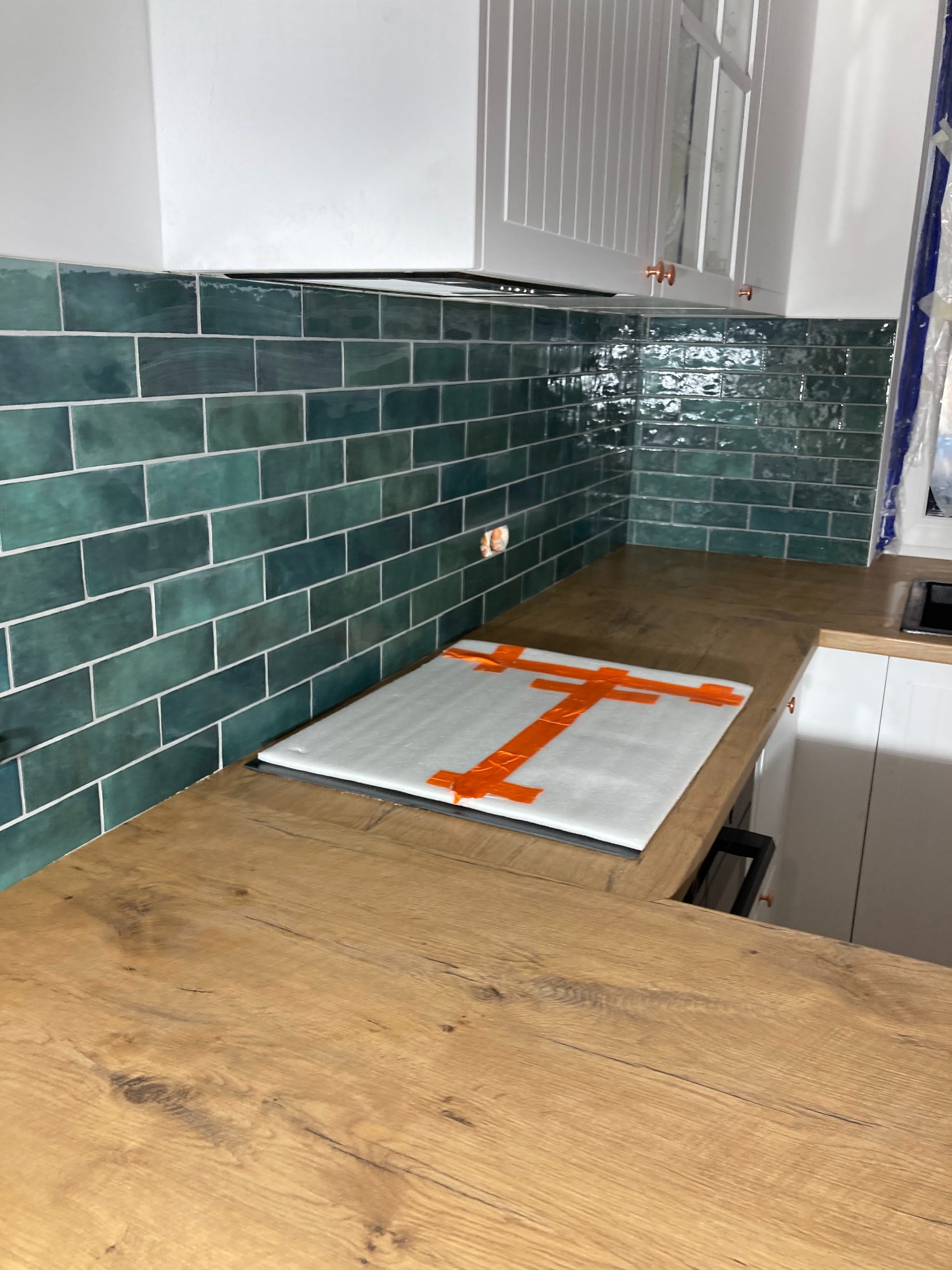 Nowoczesna kuchnia z błyszczącymi, szmaragdowymi płytkami na ścianie nad blatem, drewniany blat roboczy, białe szafki górne z miedzianymi uchwytami, płyta indukcyjna zabezpieczona folią ochronną.