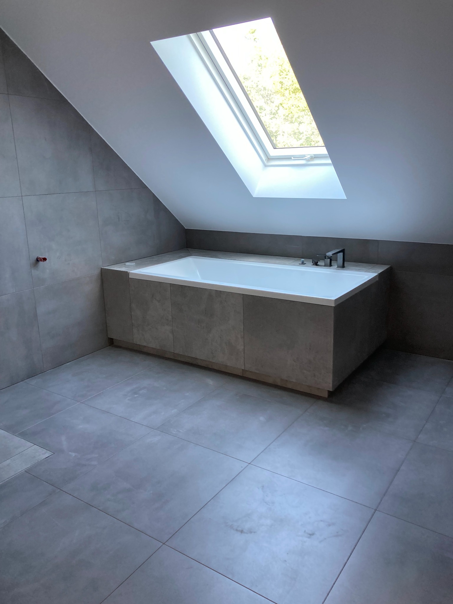 Nowoczesna łazienka na poddaszu z szarymi płytkami, białą wanną obudowaną płytkami i oknem dachowym wpuszczającym naturalne światło.