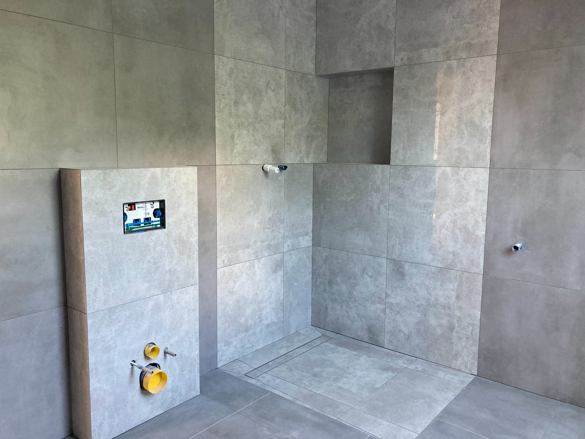 Szara łazienka w trakcie wykończenia: widoczne płytki imitujące beton, instalacja podtynkowa WC z widocznymi przyłączami oraz odpływ liniowy w podłodze.