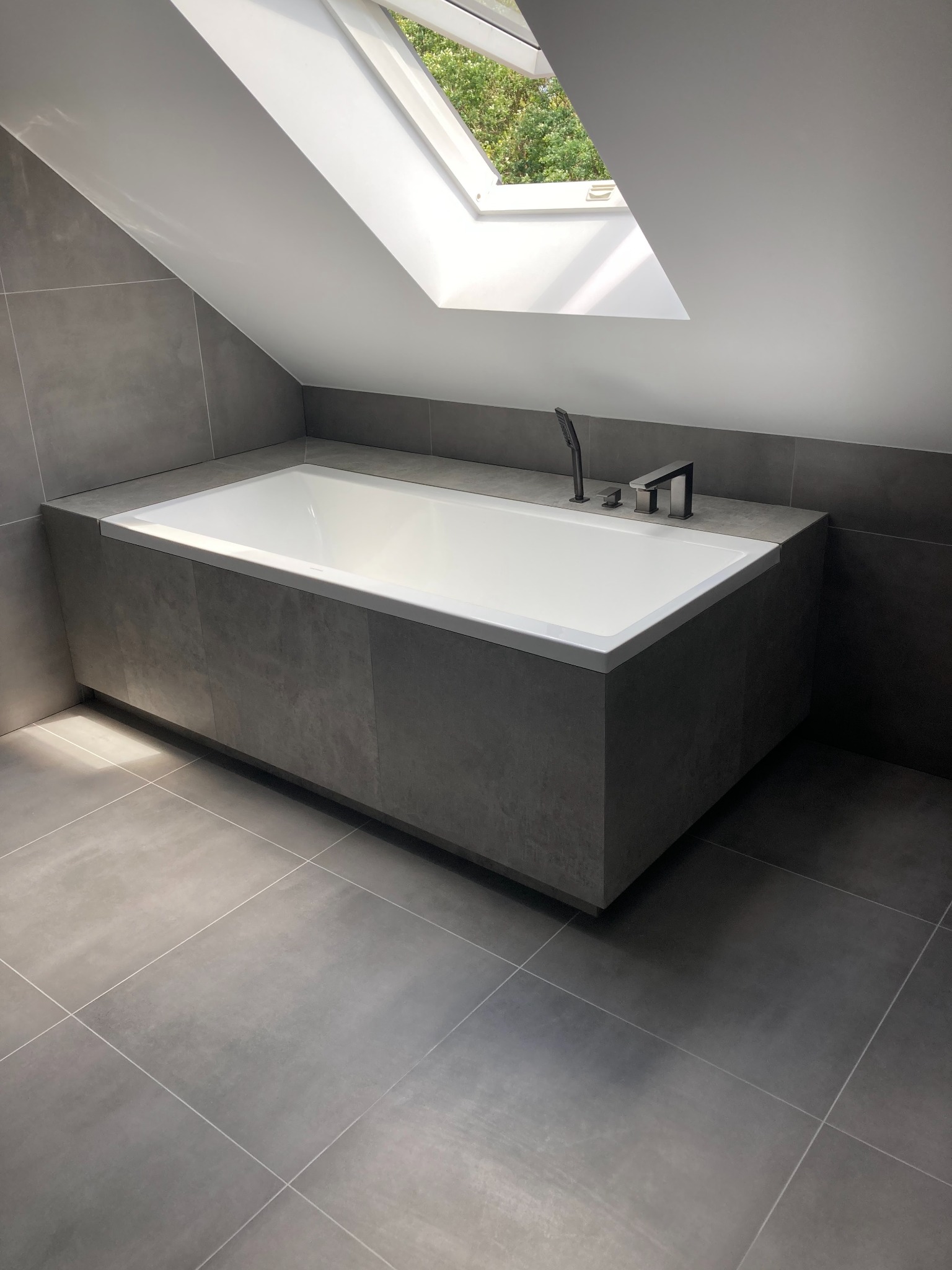 Nowoczesna łazienka na poddaszu z oknem dachowym, szara glazura na podłodze i obudowie wanny, minimalistyczna bateria wannowa w kolorze czarnym.