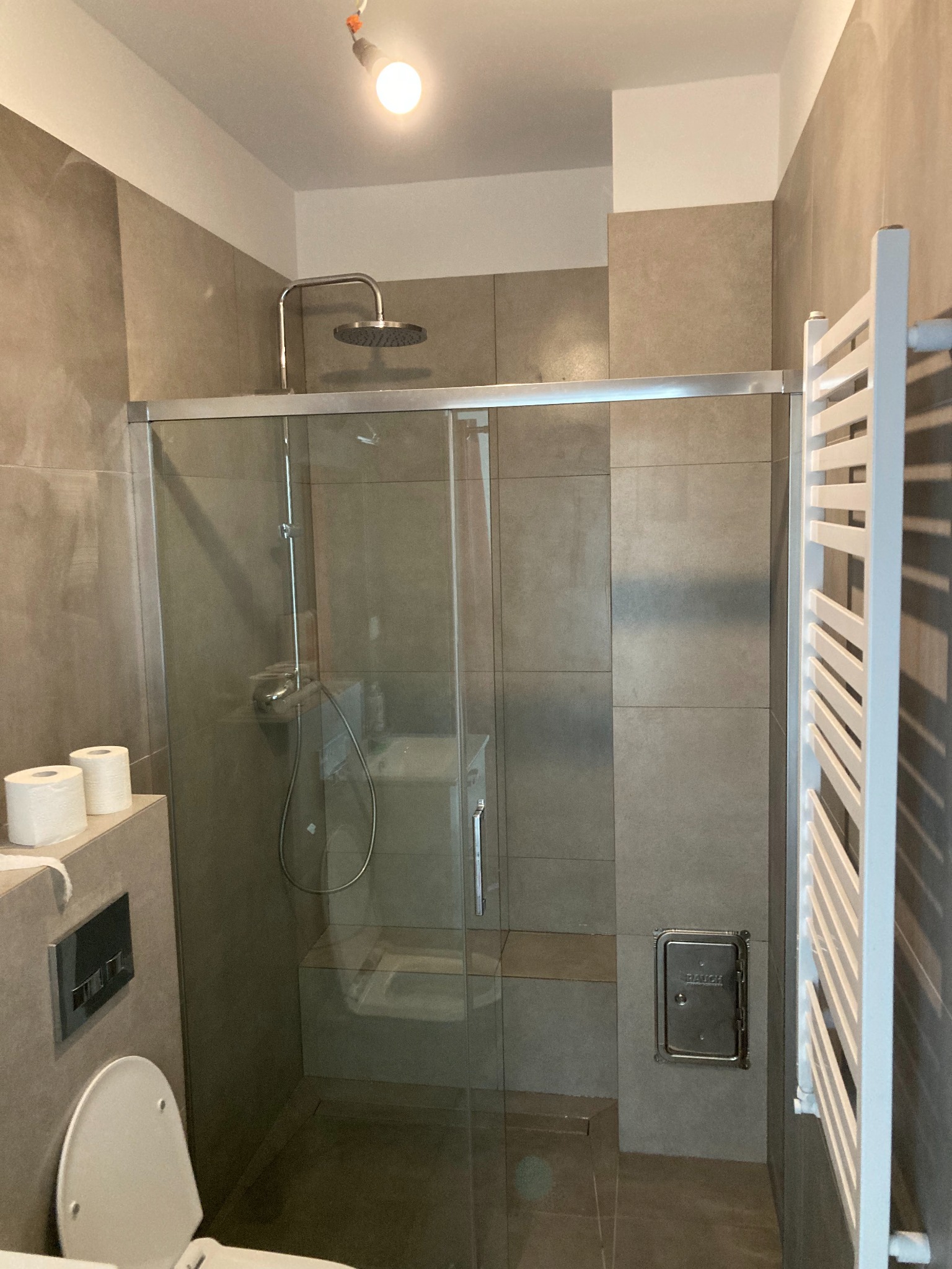 Nowoczesna łazienka z kabiną prysznicową typu walk-in, grzejnikiem drabinkowym i podwieszaną toaletą w odcieniach szarości, z widocznym włazem rewizyjnym.