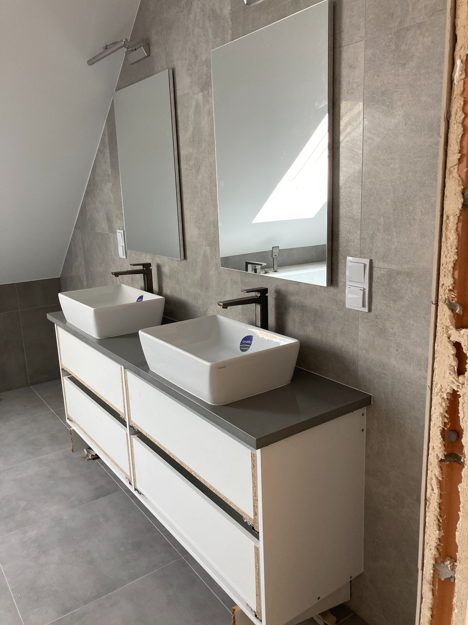 Nowoczesna łazienka w szarościach z dwoma kwadratowymi umywalkami nablatowymi, czarną armaturą i dużymi lustrami, widoczny fragment niezakończonej ściany z cegły.