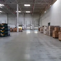 Przestronne wnętrze magazynu z betonową podłogą, paletami z towarem po lewej stronie i rzędami zapakowanych kartonów po prawej, w tle widoczne bramy dokowe i wózek widłowy.