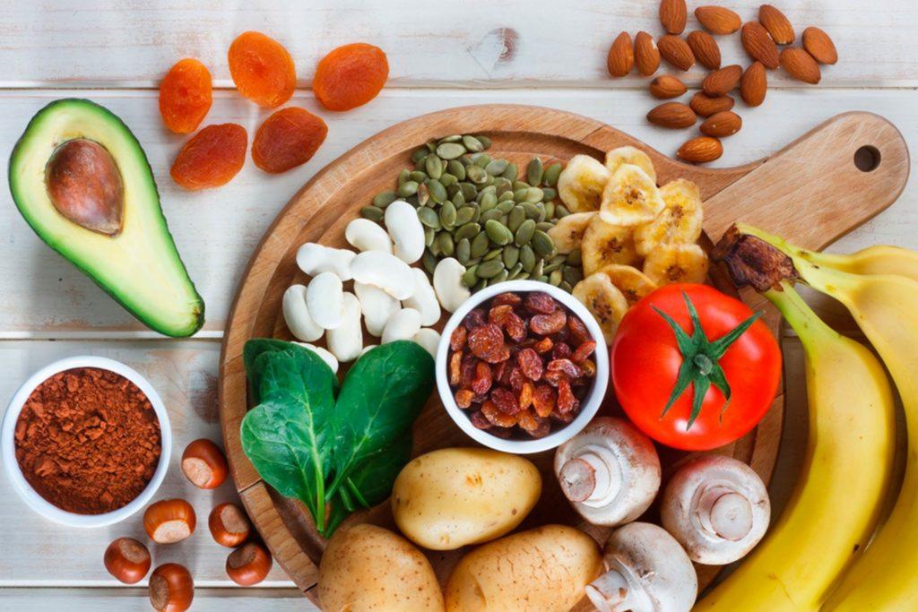 Drewniana deska z awokado, suszonymi morelami, migdałami, pestkami dyni, suszonymi bananami, rodzynkami, pomidorem, szpinakiem, ziemniakami, pieczarkami, bananami, kakao i orzechami laskowymi...