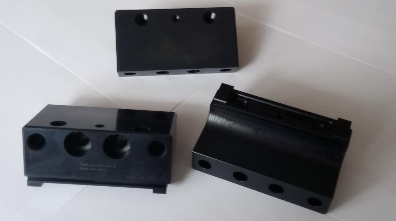 Trzy czarne, precyzyjnie obrobione elementy z tworzywa sztucznego lub metalu z otworami, leżące na jasnej powierzchni, widoczne ślady obróbki.