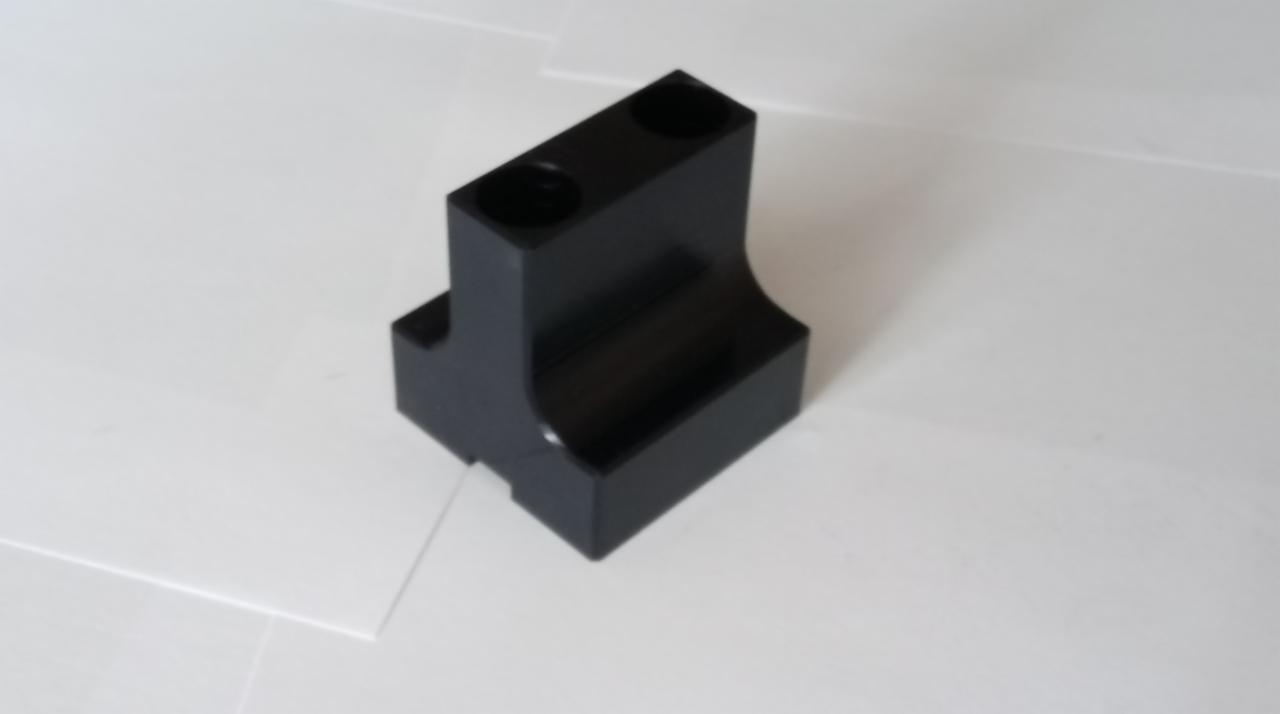 Czarny, precyzyjnie obrobiony element z dwoma otworami, widoczne ślady obróbki na krawędziach, leżący na białym tle.