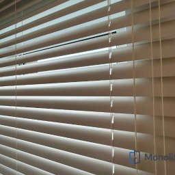 MONOLIT - Zamknięte, kremowe żaluzje poziome z widocznym logotypem producenta w prawym dolnym rogu.