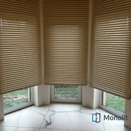 MONOLIT - Beżowe żaluzje poziome zamontowane w oknie wykuszowym, widok na zewnątrz z zielonym trawnikiem, marmurowa podłoga z szarymi żyłami, logo firmy w prawym dolnym rogu.