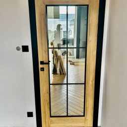 MONOLIT - Nowoczesne drzwi wewnętrzne z drewnianą ramą i czarnymi elementami, przeszklone z widocznym odbiciem sylwetki w szybie, na tle parkietu ułożonego w jodełkę.