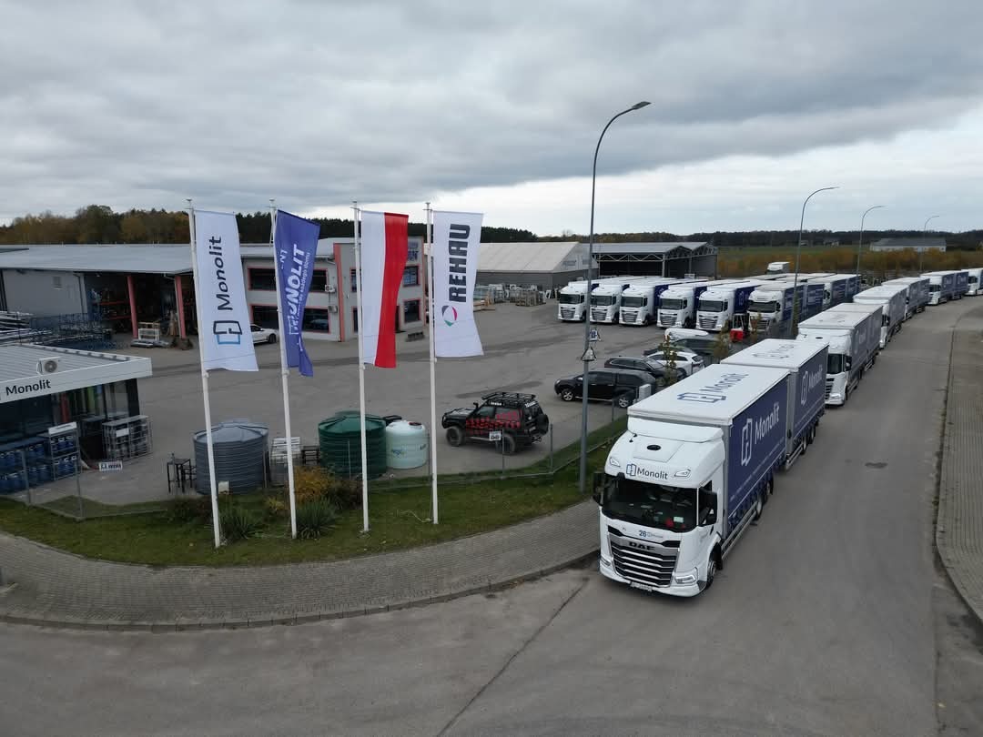 Flaga Monolit i Rehau przed budynkiem firmy, w tle zaparkowane ciężarówki Monolit czekające na załadunek.