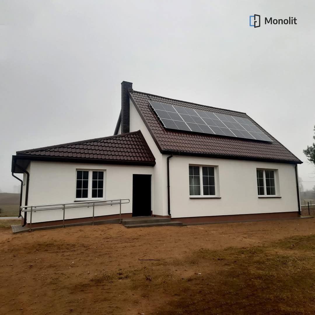 Dom jednorodzinny z białymi ścianami, brązowym dachem z panelami słonecznymi, widoczne okna z białymi ramami oraz podjazd dla niepełnosprawnych. Na dachu zamontowane panele fotowoltaiczne.