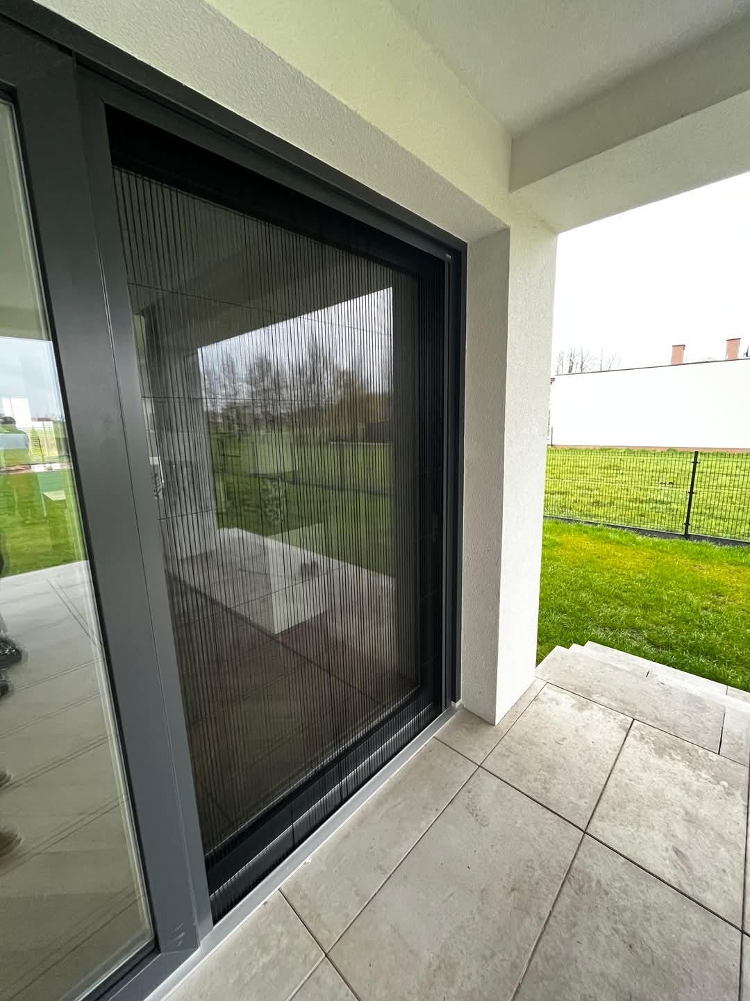 Szara rama drzwi przesuwnych z moskitierą plisowaną na tarasie wyłożonym płytkami, widok na ogród.