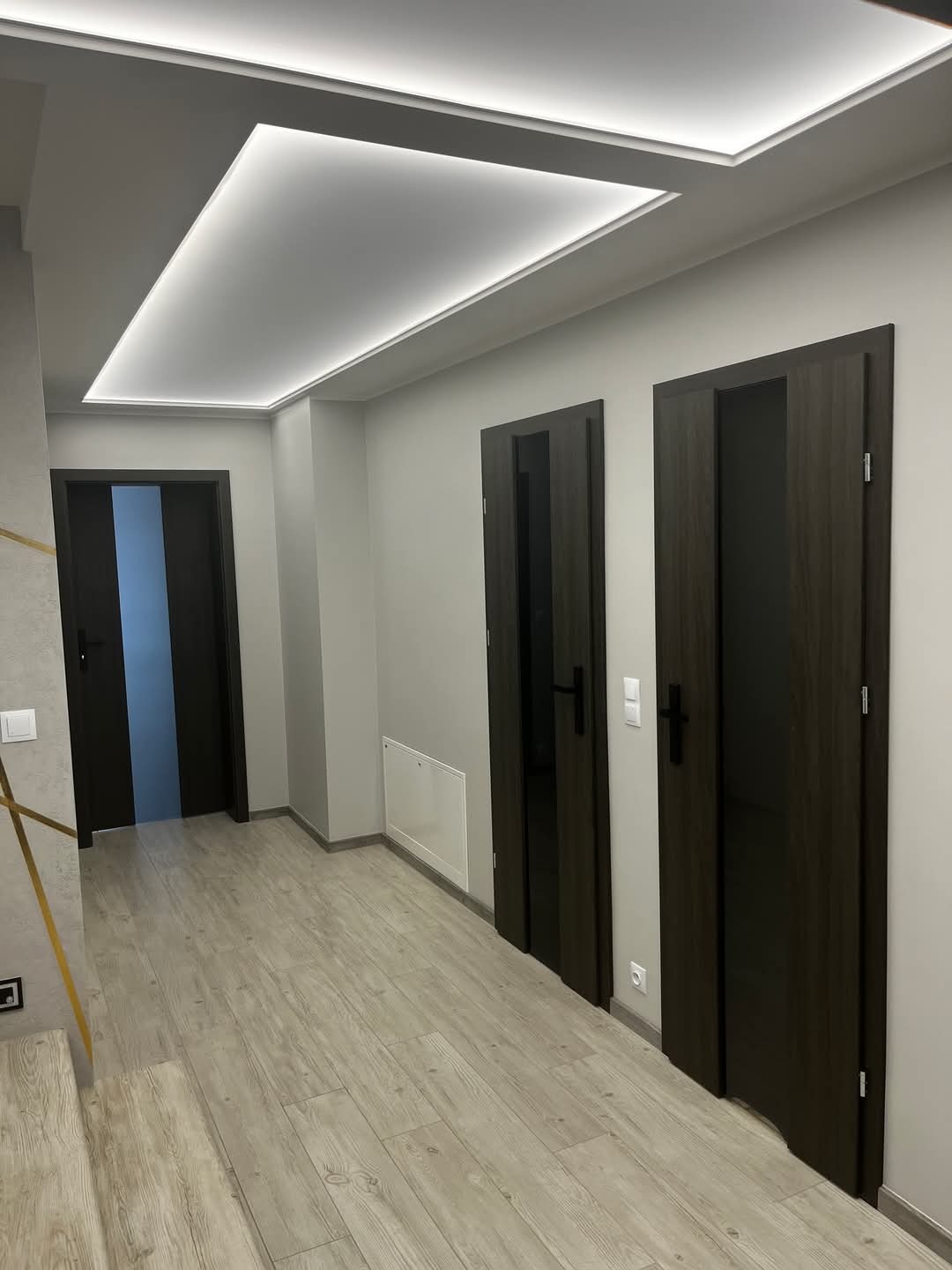 Wnętrze korytarza z trzema ciemnymi drzwiami wewnętrznymi, jasną podłogą imitującą drewno i podwieszanym sufitem z oświetleniem LED.