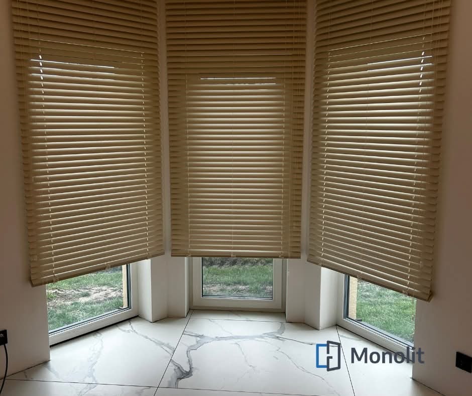 Beżowe żaluzje poziome zamontowane w oknie wykuszowym, widok na zewnątrz z zielonym trawnikiem, marmurowa podłoga z szarymi żyłami, logo firmy w prawym dolnym rogu.