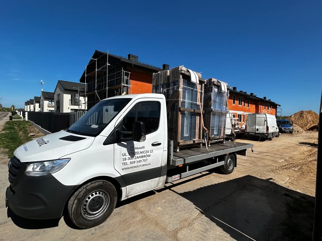 Dostawczy samochód z logo firmy Monolit przewozi zabezpieczone folią palety z oknami na plac budowy. W tle dom w stanie surowym z rusztowaniami.