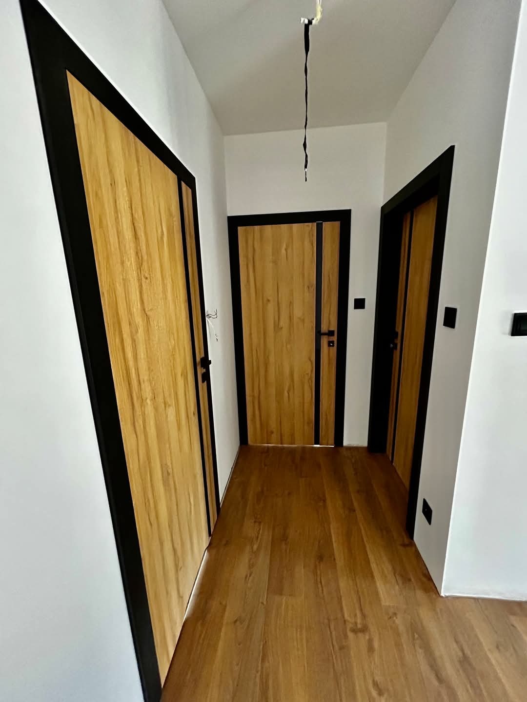 Korytarz z trzema drewnopodobnymi drzwiami z czarnymi ościeżnicami, widok na podłogę z paneli w kolorze drewna, niedokończone przewody zwisające z sufitu.