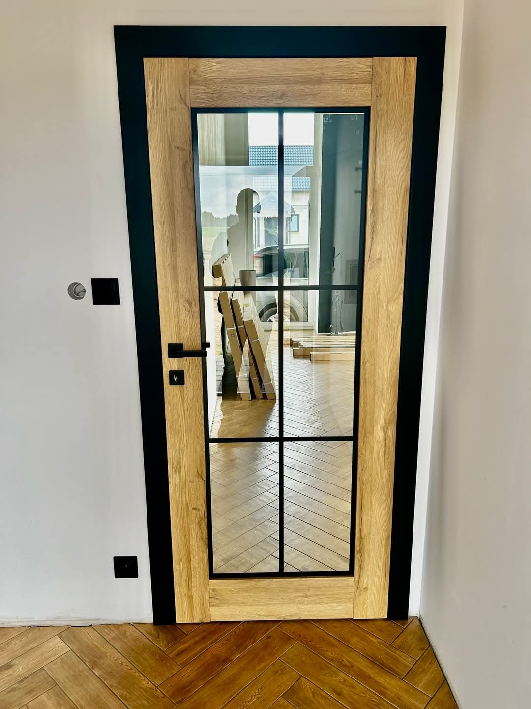 Nowoczesne drzwi wewnętrzne z drewnianą ramą i czarnymi elementami, przeszklone z widocznym odbiciem sylwetki w szybie, na tle parkietu ułożonego w jodełkę.