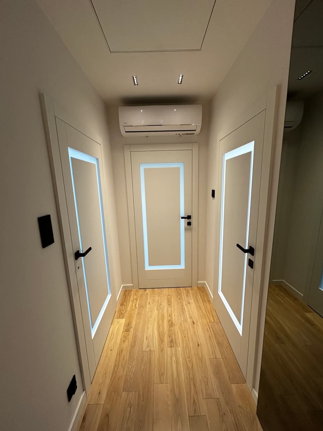 Korytarz z trzema białymi drzwiami wewnętrznymi z podświetlanymi ramkami, czarne klamki, parkiet na podłodze, klimatyzator na ścianie.