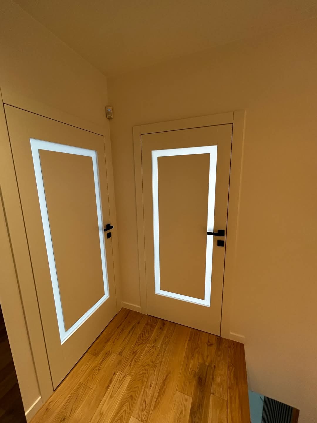 Dwa skrzydła drzwiowe wewnętrzne z podświetlanymi, prostokątnymi ramkami, zamontowane w narożniku korytarza z drewnianą podłogą i systemem alarmowym na ścianie.