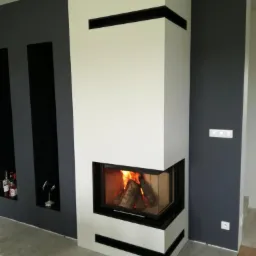 Nowoczesny narożny wkład kominkowy z szybą, obudowany białą konstrukcją z czarnymi akcentami, płomienie widoczne wewnątrz, na tle szarej ściany z wnękami.