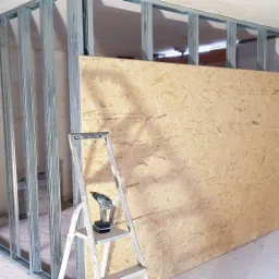 Konstrukcja ścianki działowej z profili stalowych i płyty OSB w surowym wnętrzu, w tle widoczny otw&oacute;r drzwiowy, na drabinie akumulatorowa wiertarko-wkrętarka.
