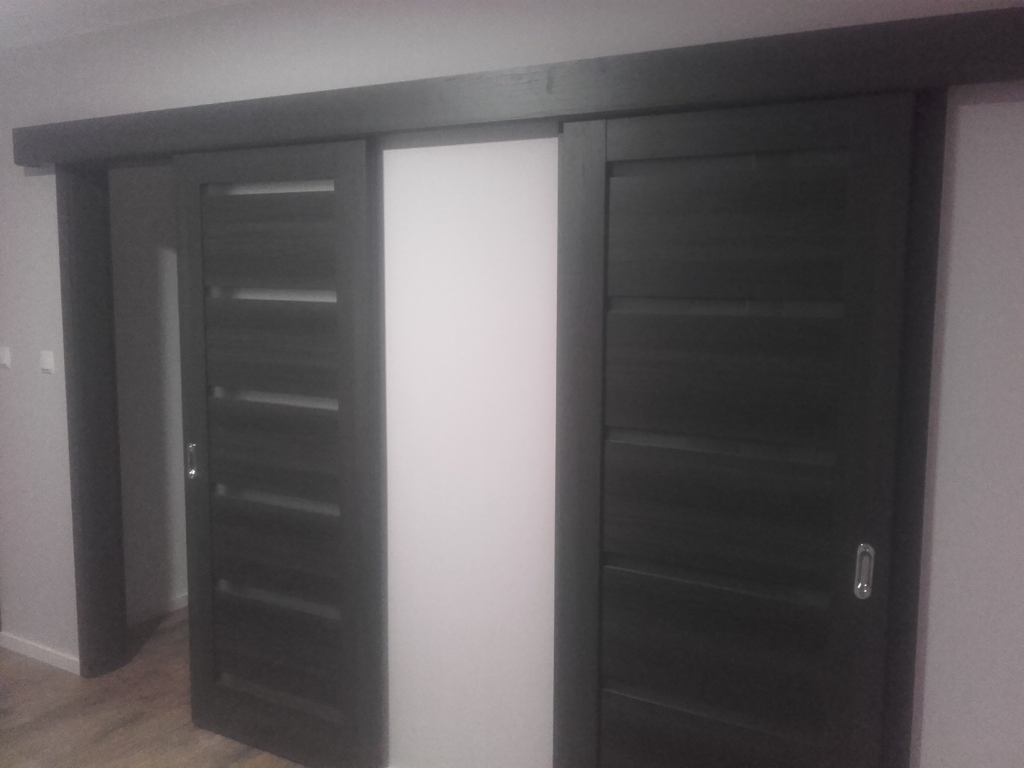 Ciemne, przesuwne drzwi z poziomymi przeszkleniami, częściowo otwarte, na tle jasnej ściany.