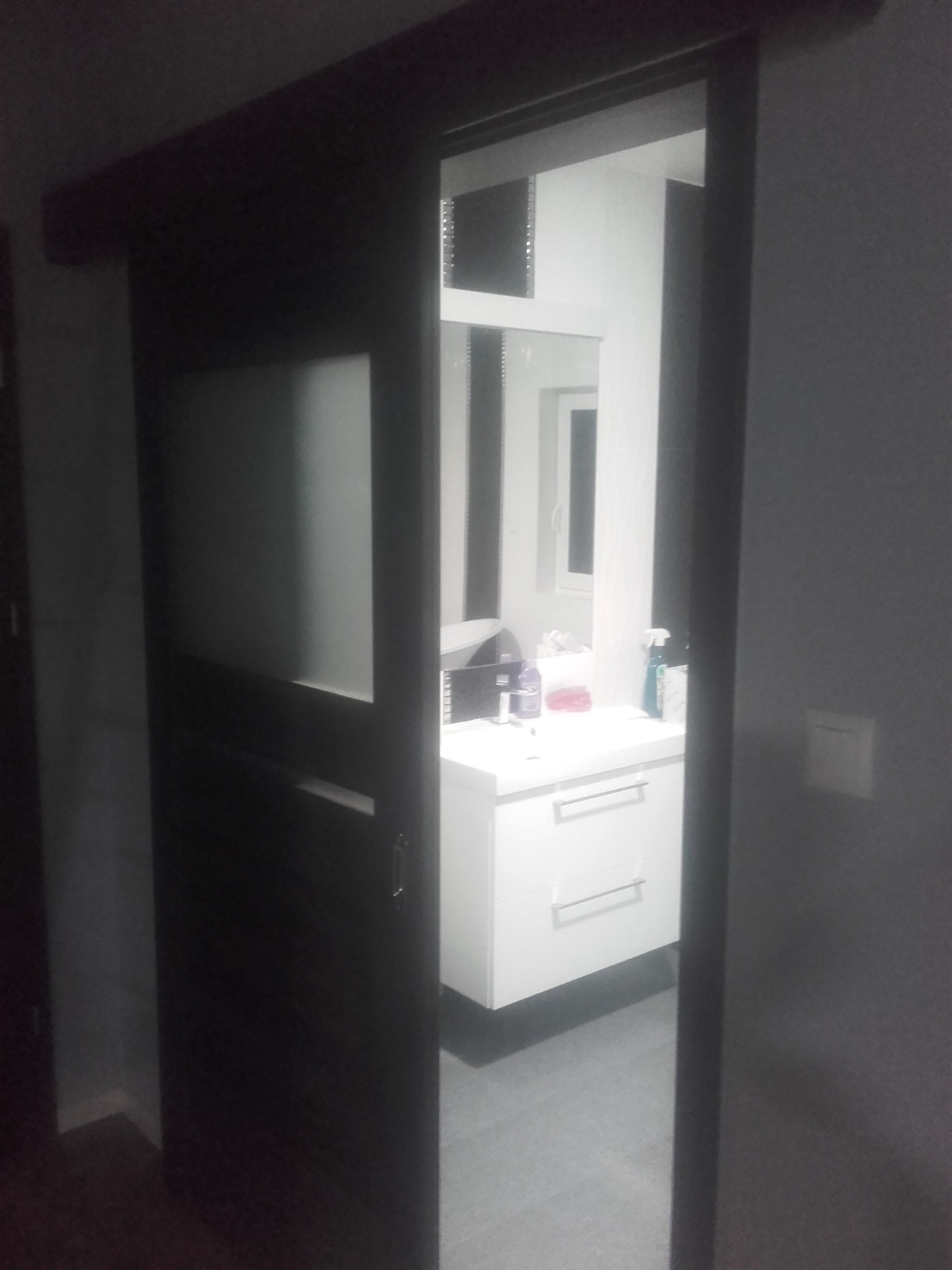 Ciemne drzwi przesuwne z matowym szkłem, uchylone, ukazujące białą szafkę podumywalkową z dwiema szufladami i blatem w łazience, widoczne akcesoria i okno w tle.