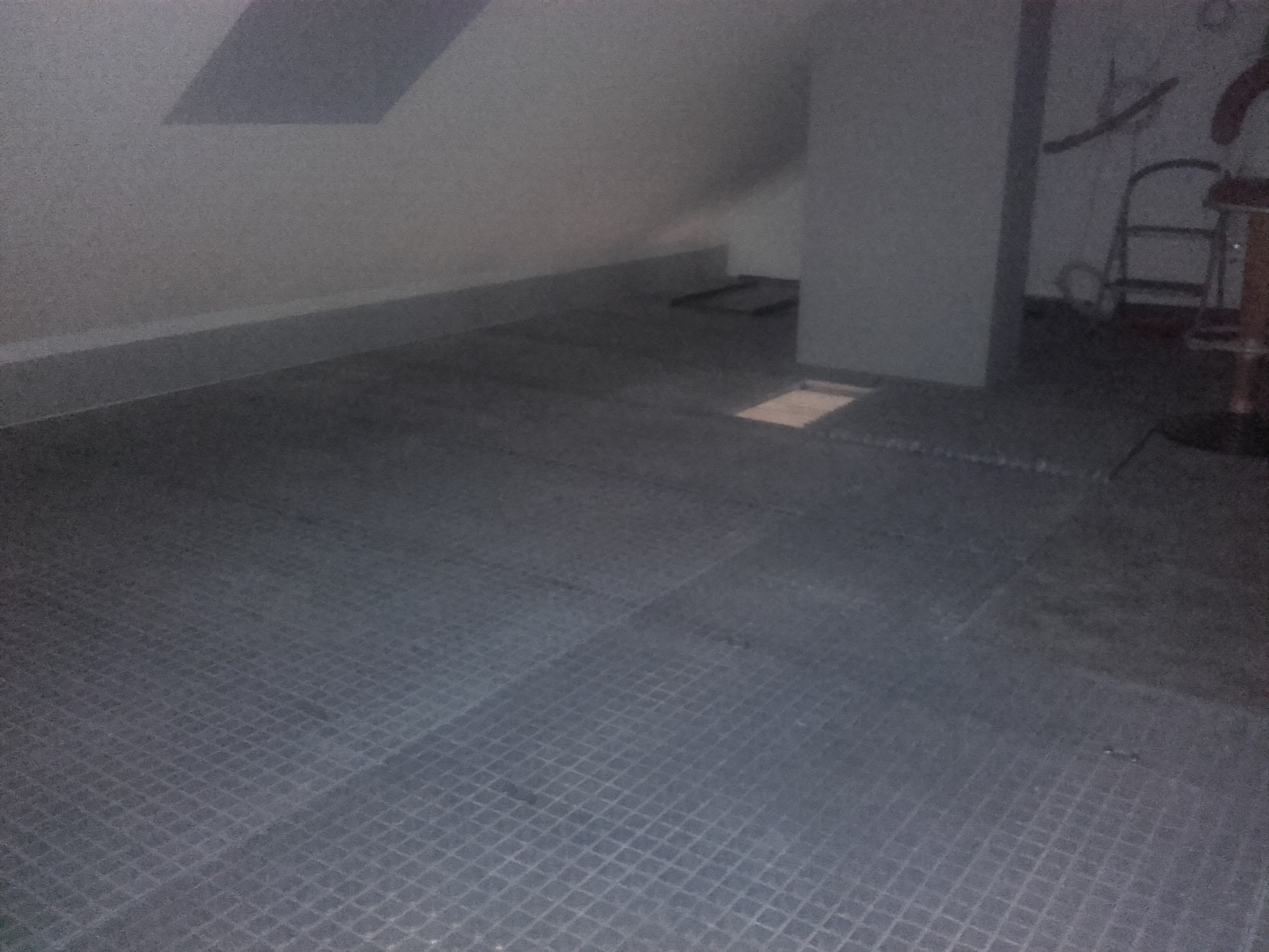 Podłoga w szarym kolorze z wzorem kratki w poddaszu, widoczny fragment ściany i kolumna, słabe oświetlenie.