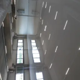 Błyszcząca, szara posadzka w hali przemysłowej, widoczne okna i instalacja wentylacyjna pod sufitem.