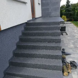 Zewnętrzne schody wejściowe do domu pokryte szarą, chropowatą nawierzchnią, z widocznymi śladami prac wykończeniowych i drewnem zabezpieczonym folią obok.