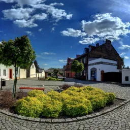 Ujęcie miejskiego placu z budynkami w tle, w tym budynku z napisem 'Skuteczne strony www' nad wejściem, oświetlonego słońcem i błękitnym niebem z chmurami.