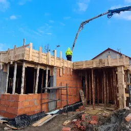 Budowa domu jednorodzinnego w trakcie murowania ścian z czerwonej cegły, widoczne drewniane szalunki, pracownik w kamizelce odblaskowej oraz ramię pompy do betonu na tle błękitnego nieba.