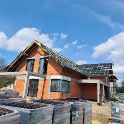 Dom murowany w stanie surowym otwartym, widoczne palety z dachówką i konstrukcja dachu, błękitne niebo w tle.