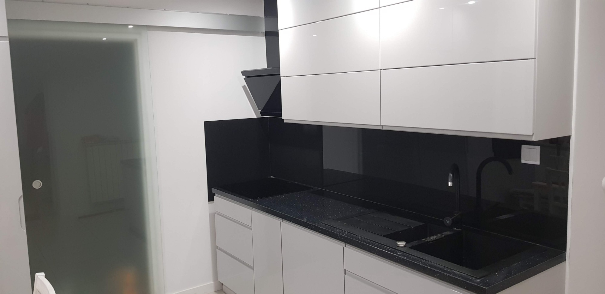 Nowoczesna kuchnia z białymi, błyszczącymi szafkami, czarnym blatem z brokatem i czarnym panelem szklanym nad blatem, widoczna również matowa szklana przesuwana ściana.
