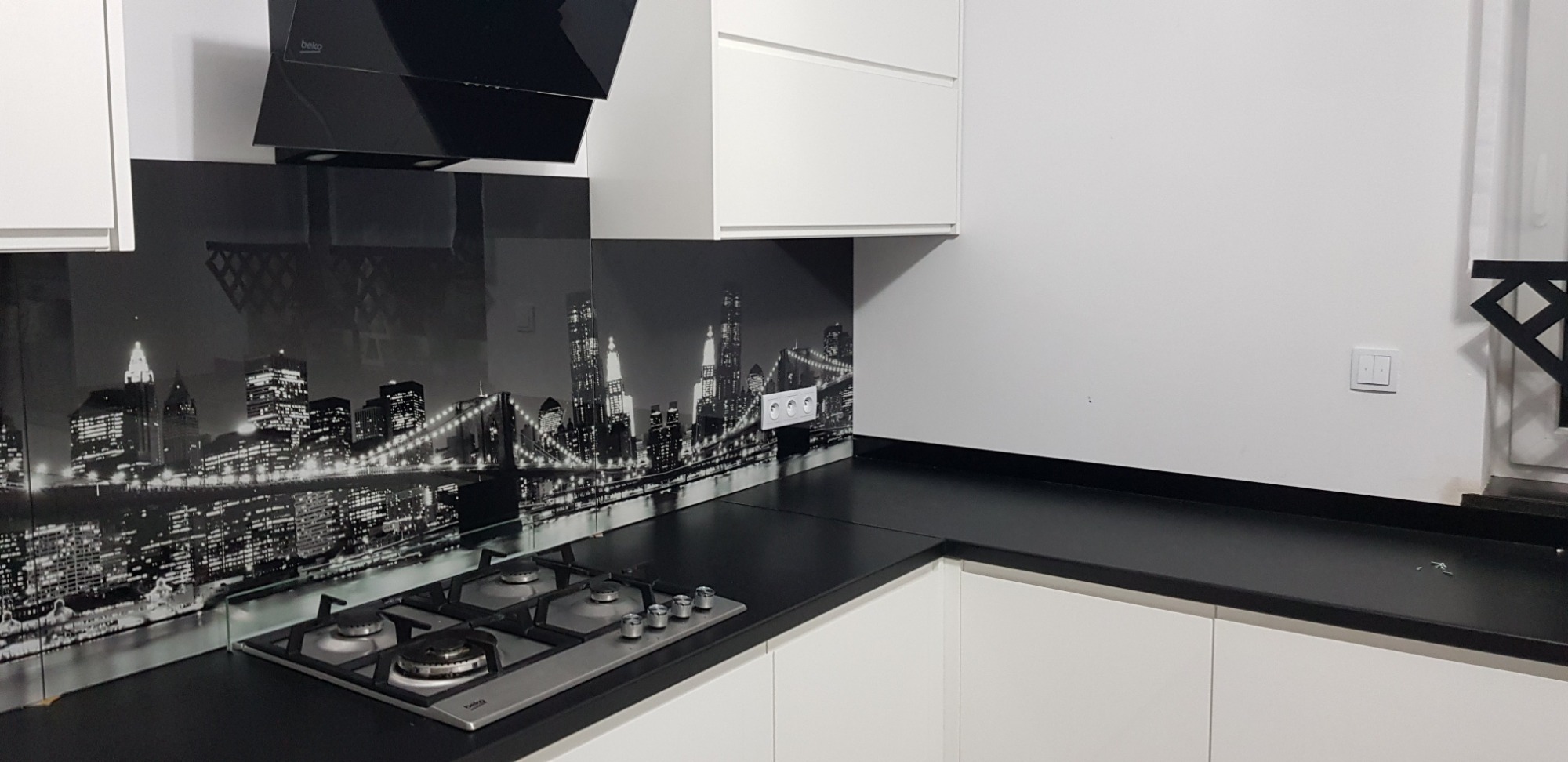 Szklany panel kuchenny z czarno-białym motywem panoramy Nowego Jorku, zamontowany nad płytą gazową w nowoczesnej kuchni z białymi szafkami i czarnym blatem.