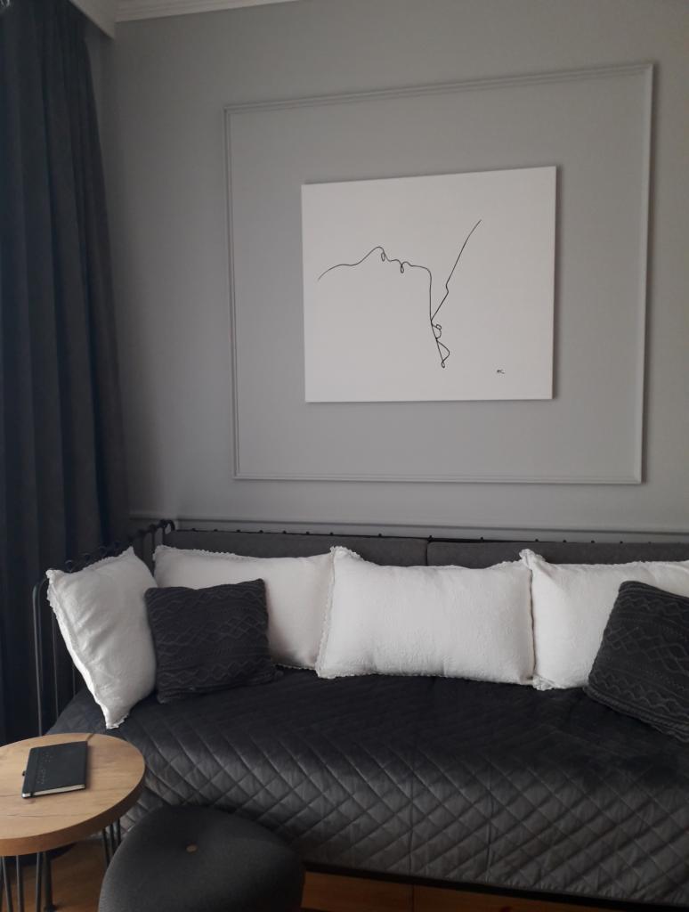 Szary pokój z czarną sofą, białymi i szarymi poduszkami, okrągłym stolikiem z notatnikiem i minimalistyczną grafiką na ścianie w ozdobnej ramie.