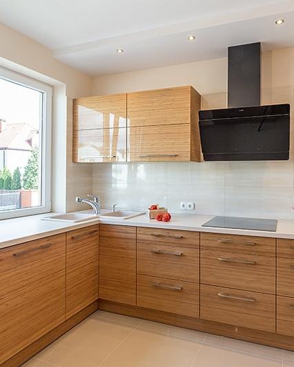 Jasna kuchnia z drewnianymi szafkami o poziomym usłojeniu, czarnym okapem i białym blatem, uzupełniona o świeże pomidory na blacie.