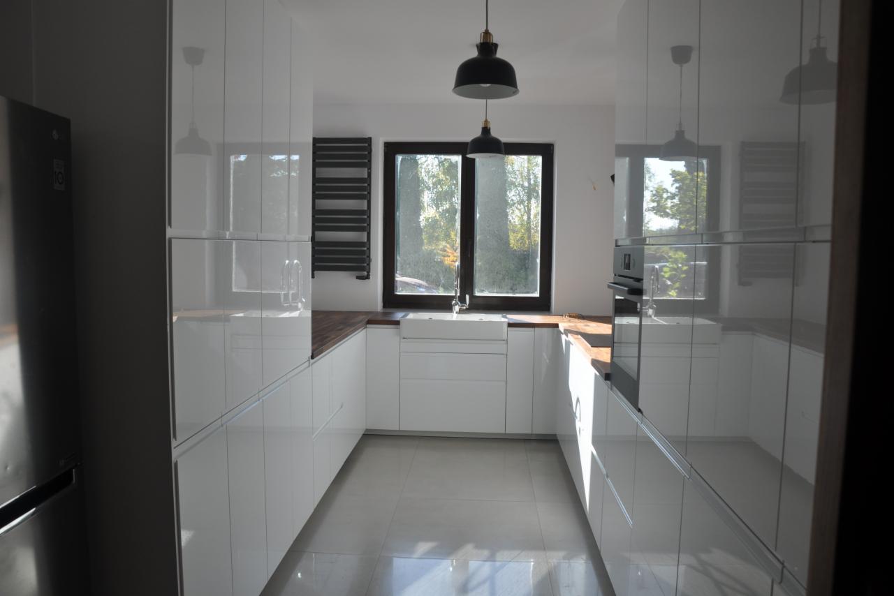 Nowoczesna, biała kuchnia z drewnianym blatem w mieszkaniu w Warszawie, z dużym oknem i czarnymi lampami wiszącymi, widziana z perspektywy wejścia.