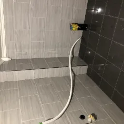 Nowa łazienka w trakcie wykańczania: jasnoszare płytki imitujące drewno na ścianach i podłodze, widoczny odpływ liniowy, czarna rura wystająca ze ściany z podłączonym białym wężem, fragment listwy...