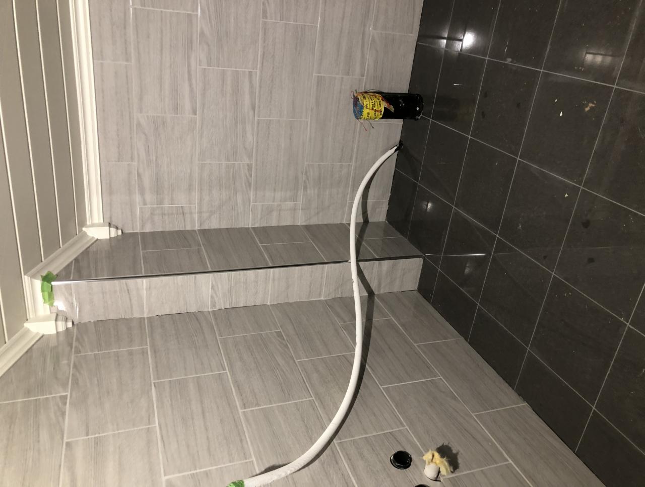 Nowa łazienka w trakcie wykańczania: jasnoszare płytki imitujące drewno na ścianach i podłodze, widoczny odpływ liniowy, czarna rura wystająca ze ściany z podłączonym białym wężem, fragment listwy...