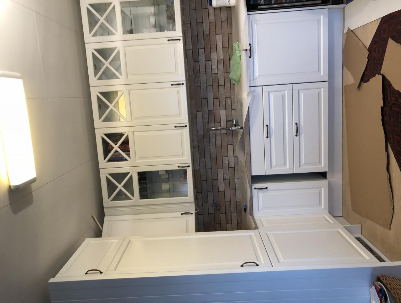 Biała zabudowa kuchenna z przeszklonymi szafkami górnymi i ciemnymi uchwytami, ściana wykończona cegiełką, zabezpieczona folią malarską, widoczne ślady prac wykończeniowych.