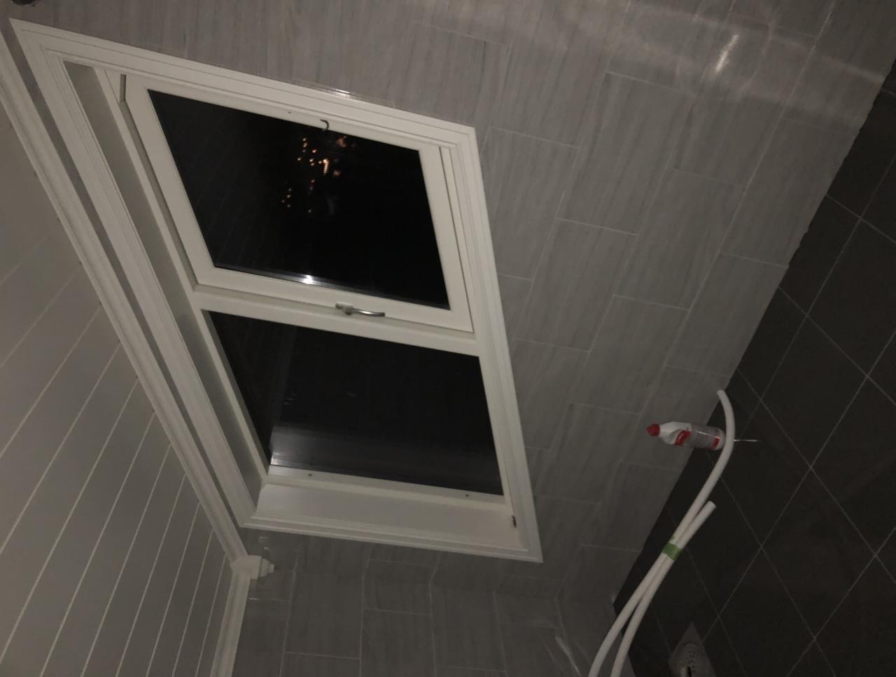 Okno dachowe z białymi ramami w łazience wyłożonej szarymi płytkami, widoczne rury i butelka po napoju.