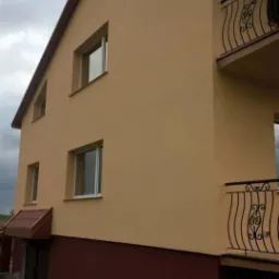 Dwupiętrowy dom z beżową elewacją, ciemnoczerwoną podmurówką i balkonami z kutymi balustradami, sfotografowany w pochmurny dzień.